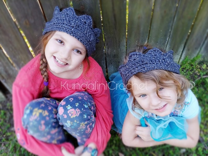May include: Two young girls wearing gray crocheted crown headbands. The girl on the left is wearing a pink sweater and blue jeans with a floral pattern. The girl on the right is wearing a blue tulle dress.