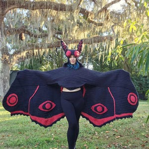 May include: A black and red crochet moth costume with large wings and a matching headpiece. The wings feature red eye-like designs and are spread wide. The headpiece has antennae and large red eye-like details.