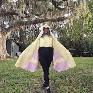 May include: A yellow crochet hooded cape with pink accents. The cape has long, wing-like sleeves and a hood with pointed ears. The person is wearing black leggings and shoes. The background features a grassy area and large trees.