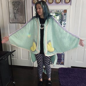 May include: A handmade, mint green crochet cape with a hood, featuring yellow and gray accents. The cape has a light purple trim and a cream-colored border. The design includes two yellow, boot-shaped pockets. The cape is open in the front and has a flowing, wing-like shape.