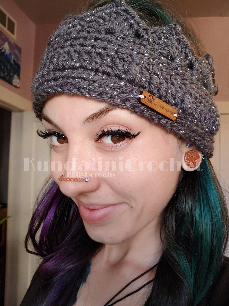 May include: A gray crocheted headband with silver glitter, featuring a crown design and a brown leather tag with the text "Kundalini Crochet" in white lettering. The headband is worn by a person with purple and teal hair, and they are wearing a nose ring with multiple beads.