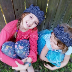 May include: Two young girls wearing gray crocheted crown headbands. One girl is wearing a pink sweater and floral pants, while the other is wearing a blue dress. The girls are sitting in front of a wooden fence.
