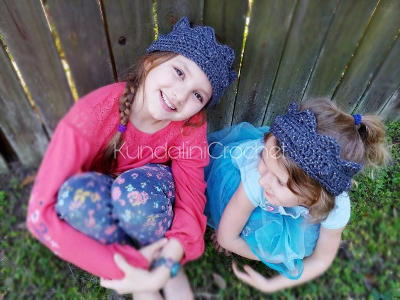 May include: Two young girls wearing gray crocheted crown headbands. One girl is wearing a pink sweater and floral pants, while the other is wearing a blue dress. The girls are sitting in front of a wooden fence.