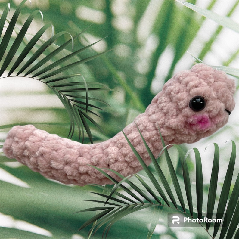 May include: A plush, light pink worm-like creature with black eyes and a pink mouth. The handmade toy is set against a blurred green and white background with palm leaves. The worm has a textured, soft appearance.