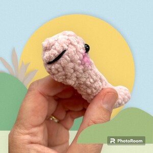May include: A handmade, light pink crocheted creature with a smiling face, black eyes, and a pink mouth. The plush toy is held in a hand against a backdrop of a yellow circle and a blue sky.