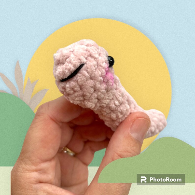 May include: A handmade, light pink crocheted creature with a smiling face, black eyes, and a pink mouth. The plush toy is held in a hand against a backdrop of a yellow circle and a blue sky.