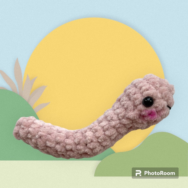 May include: A handmade, light pink crocheted worm with black safety eyes and pink cheeks. The worm is made of a soft, textured yarn and is posed against a colorful background with a yellow circle and green and blue shapes.