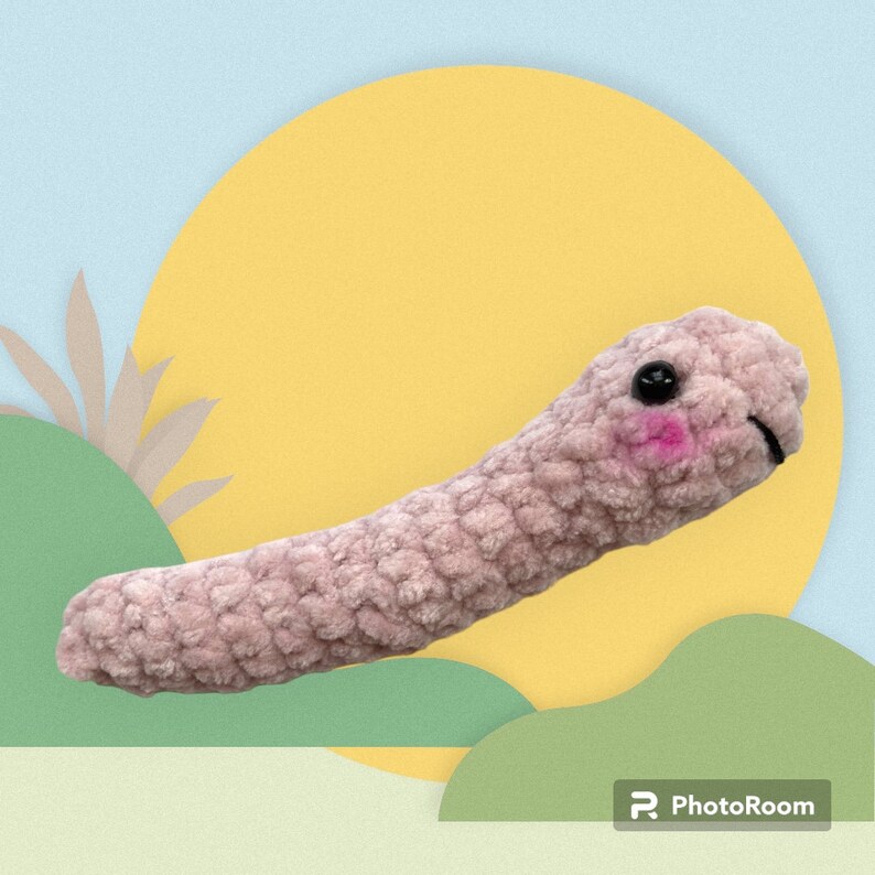 May include: A plush, pink worm-shaped toy with black button eyes and rosy cheeks. The soft, textured toy has a smiling mouth and is set against a colorful background with a yellow circle and green shapes.