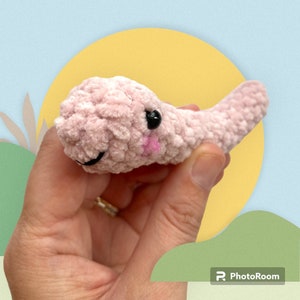 May include: A handmade, light pink crocheted creature with black eyes and a smiling mouth. The plush toy has a small pink blush on its cheeks and a tail. The toy is held in a hand against a colorful background.