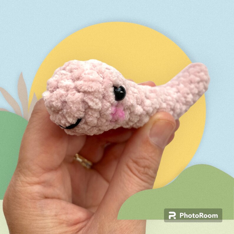 May include: A handmade, light pink crocheted creature with black eyes and a smiling mouth. The plush toy has a small pink blush on its cheeks and a tail. The toy is held in a hand against a colorful background.