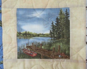 May include: A quilted fabric panel featuring a scenic image of a lake, a wooden jetty, a red rowing boat, and two green pine trees. The background is a blue sky with white clouds.