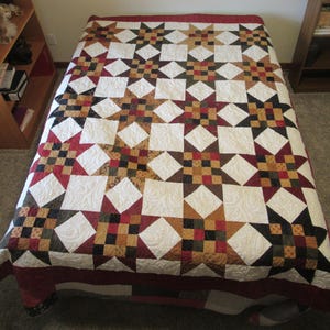 May include: A quilt with a star pattern in shades of red, brown, black, and gold. The quilt is made of patchwork squares and has a white background.