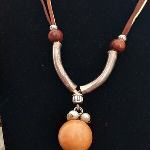 May include: A brown leather necklace with a silver pendant featuring a large orange bead and smaller white beads. The necklace is displayed on a black mannequin.