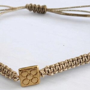 May include: A gold charm bracelet with a square charm featuring a flower design. The bracelet is made of beige cord and has adjustable closures.