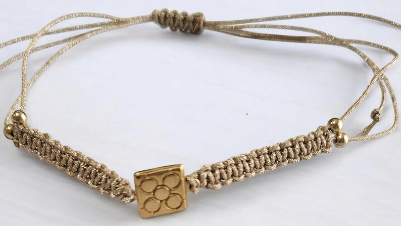 May include: A gold charm bracelet with a square charm featuring a flower design. The bracelet is made of beige cord and has adjustable closures.