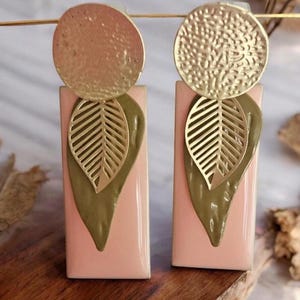 May include: A pair of gold and green statement earrings with a pink background. The earrings feature a round gold disc, a gold leaf design, and a green leaf shape.