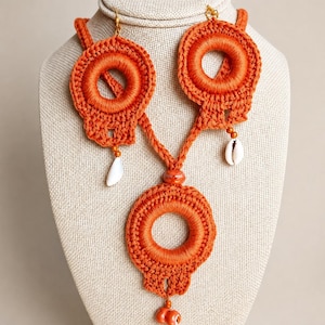 May include: A vibrant orange crochet necklace featuring three circular motifs connected by a central cord. The design includes two small white cowrie shells hanging from the upper circles and a small orange bead accent at the bottom center.