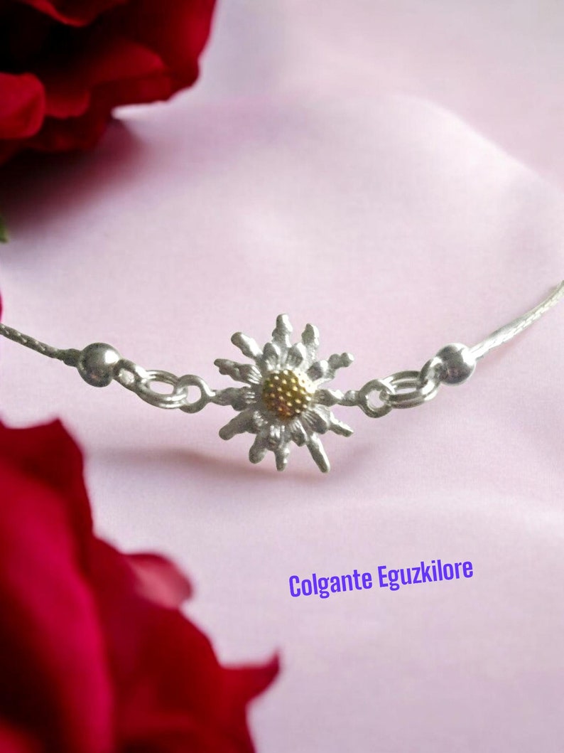 May include: A silver chain necklace with a delicate daisy charm. The daisy has a gold center and silver petals. The necklace is on a pink background with a red rose.
