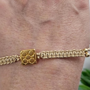 May include: A gold bracelet with a square charm featuring a geometric design. The bracelet is made of a beige cord and has gold beads.