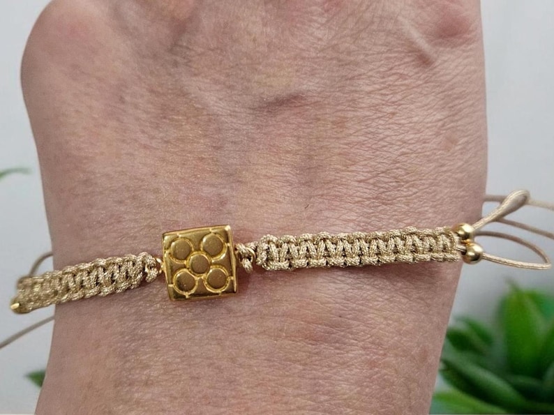 May include: A gold bracelet with a square charm featuring a geometric design. The bracelet is made of a beige cord and has gold beads.