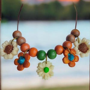 May include: A handmade necklace featuring wooden beads in brown, orange, and green, interspersed with cream-colored flower pendants with brown and blue accents. The necklace is strung on a brown cord, perfect for a bohemian look.