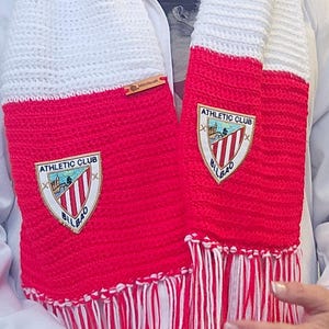 May include: A red and white knitted scarf with the "ATHLETIC CLUB BILBAO" logo. The scarf has a fringed edge and a small wooden tag that reads "Montseuk". The scarf is red with white stripes and white ends.
