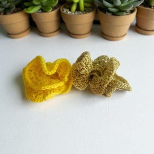 以下が含まれることがあります： 2つの手作りのかぎ針編みアイテム。1つは明るい黄色、もう1つは金色です。白い表面にあり、背景にはいくつかの小さな鉢植えの多肉植物があります。