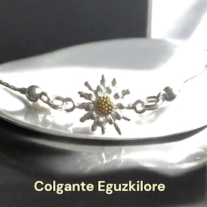 May include: A silver chain necklace with a silver and gold flower pendant. The flower has a yellow center and white petals. The necklace is on a white surface. The text "Colgante Eguzkilore" is visible below the necklace.