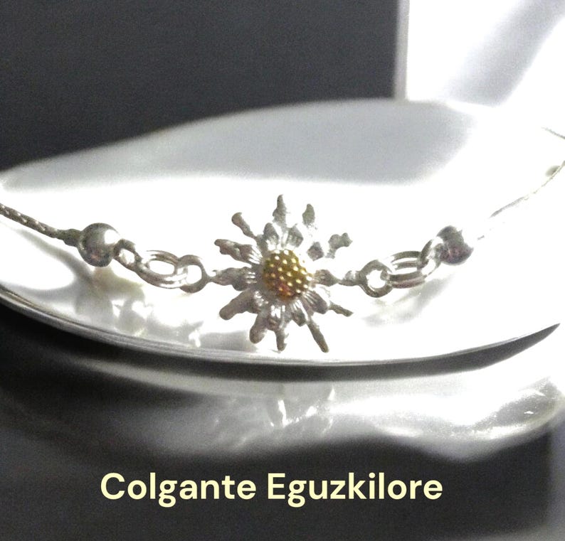 May include: A silver chain necklace with a silver and gold flower pendant. The flower has a yellow center and white petals. The necklace is on a white surface. The text "Colgante Eguzkilore" is visible below the necklace.