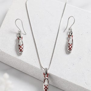 May include: A silver jewelry set consisting of a necklace and two drop earrings. Each piece features a miniature ballet slipper pendant with a textured silver finish, accented by vibrant red enamel detailing on the laces and ribbon ties.