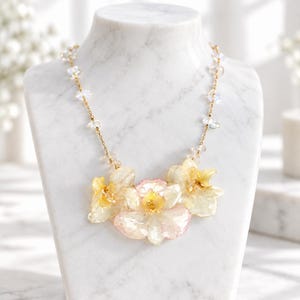 May include: A necklace featuring three pressed, translucent yellow and pink orchid flowers as a central pendant. The chain is made of thin gold-tone metal links interspersed with small, clear, iridescent crystal beads along the upper sections.