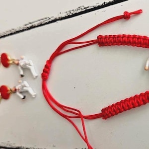 May include: Red string bracelet with a miniature figure in a white outfit. The bracelet features a braided design and small gold-colored charms. The background is a white wooden surface.