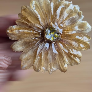 May include: A flower-shaped brooch with a glossy, resin-like finish. The petals are a light tan and brown color with a textured appearance. The center of the flower features a cluster of small, sparkling gold and yellow flecks. The word Broche is visible.