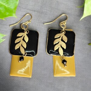May include: A pair of gold earrings with black and yellow square-shaped charms. Each charm features a gold leaf design and a black bead.