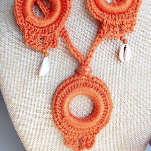 May include: Orange crochet necklace and earrings set. The necklace features a large circular pendant and is connected to two circular earrings. Each piece has a unique design with shell and bead accents. The jewelry is displayed on a mannequin.