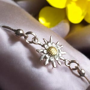 May include: A silver bracelet with a gold-toned edelweiss flower charm. The bracelet is made of a delicate chain and has a simple design.