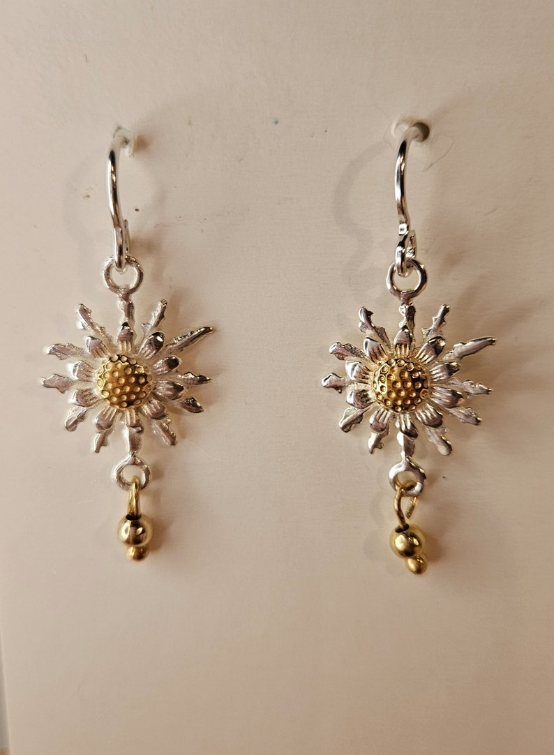 Op de afbeelding: Een paar zilverkleurige oorbellen met een gestructureerd, stervormig bloemmotief en een goudkleurig middenstuk. Elke oorbel hangt aan een zilverkleurige haak en heeft onderaan een kleine, bungelende goudkleurige kraal.