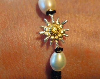 Op de afbeelding: Een zwarte gevlochten armband met twee witte parels en een gouden bloembedel.