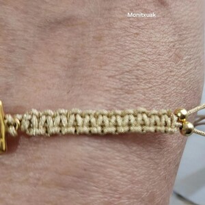 May include: A beige braided bracelet with a gold square charm and two gold beads. The bracelet has a sliding knot closure.
