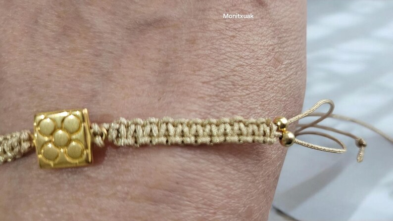 May include: A beige braided bracelet with a gold square charm and two gold beads. The bracelet has a sliding knot closure.