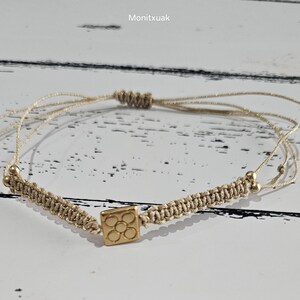 May include: A beige string bracelet with a gold charm featuring a square design with multiple circles inside. The bracelet is tied with a knot and has small gold beads on each end.