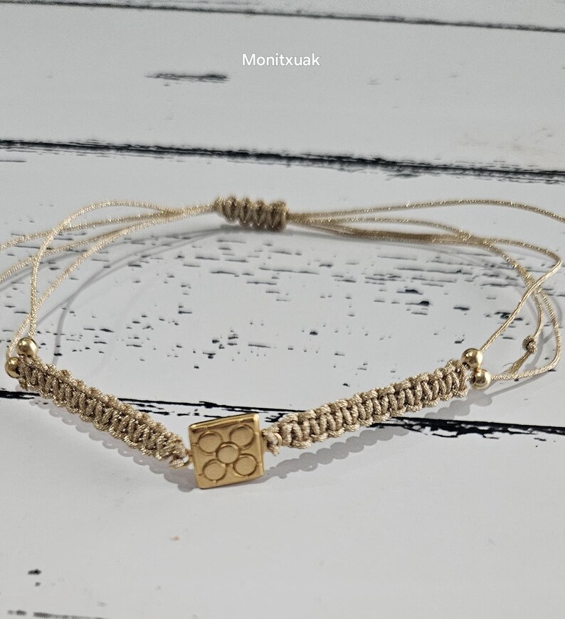 May include: A beige string bracelet with a gold charm featuring a square design with multiple circles inside. The bracelet is tied with a knot and has small gold beads on each end.