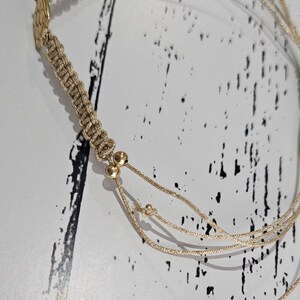 May include: A beige macrame bracelet with gold beads and a gold charm. The bracelet is made of multiple strands of cord and has a knotted design.