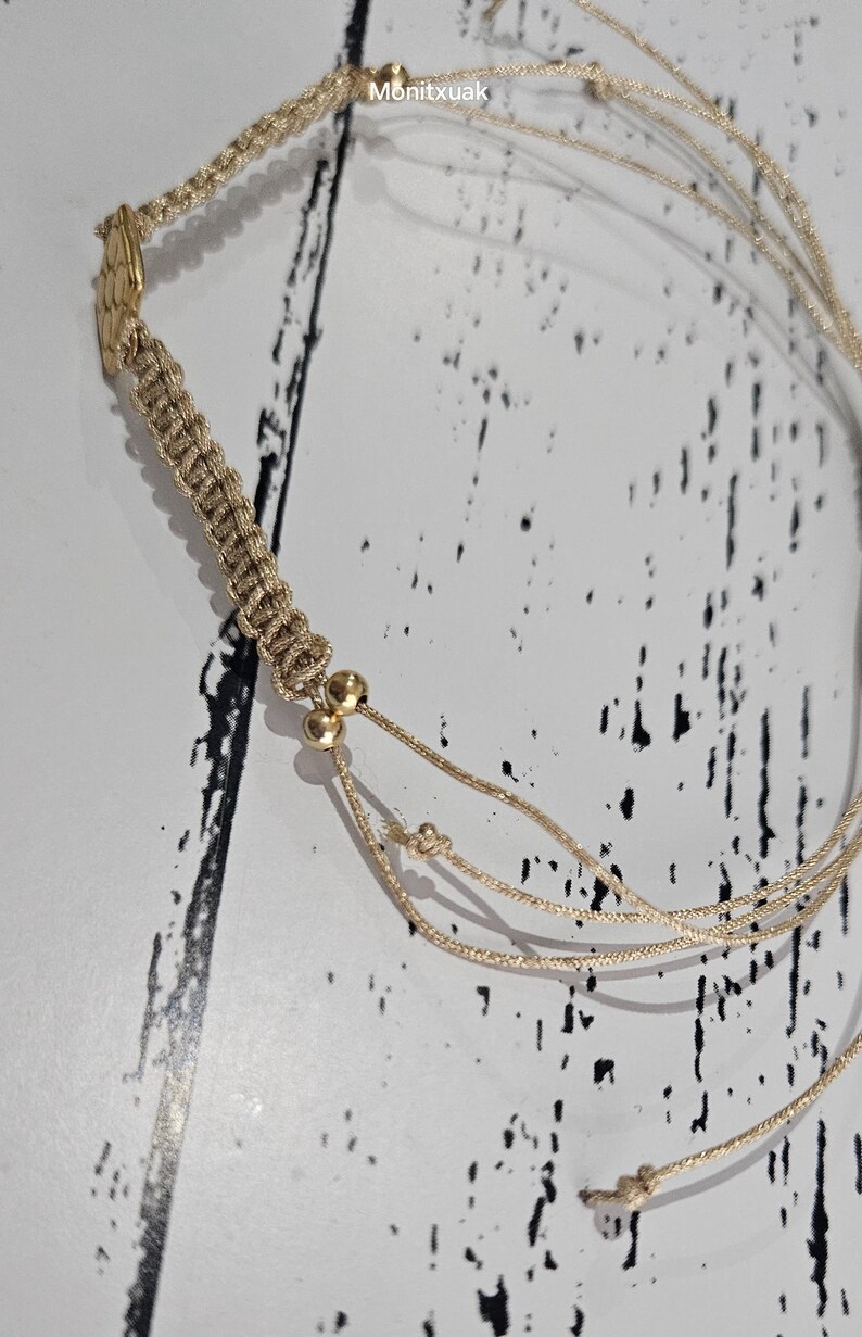 May include: A beige macrame bracelet with gold beads and a gold charm. The bracelet is made of multiple strands of cord and has a knotted design.