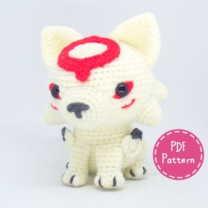 May include: White crocheted amigurumi wolf with a red circle on its forehead and black eyes. The wolf is sitting with its paws tucked under its body. The text "PDF Pattern" is on a pink circle with a dashed border.