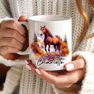 Peut inclure: Un mug en céramique blanche orné d'une illustration colorée représentant un cheval brun dans une forêt automnale aux feuillages orange et jaune. Le prénom Charlotte est inscrit en lettres cursives sombres sous le motif du cheval.