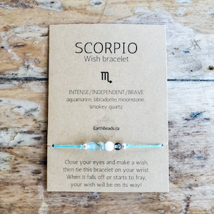 May include: A light blue string bracelet with aquamarine, labradorite, moonstone, and smokey quartz beads. The card reads "Scorpio Wish Bracelet" with the astrological symbol for Scorpio and the text "INTENSE/INDEPENDENT/BRAVE aquamarine, labradorite, moonstone, smokey quartz EarthBeads.ca Close your eyes and make a wish, then tie this bracelet on your wrist. When it falls off or starts to fray, your wish will be on its way!"