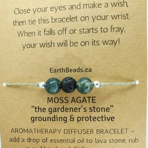 May include: A green moss agate stone bracelet with a black lava stone bead and silver spacer beads. The bracelet is tied with a light green string. The card reads "Close your eyes and make a wish, then tie this bracelet on your wrist. When it falls off or starts to fray, your wish will be on its way!"  "EarthBeads.ca"  "MOSS AGATE"  "the gardener's stone"  "grounding & protective"  "AROMATHERAPY DIFFUSER BRACELET - add a drop of essential oil to lava stone, rub"