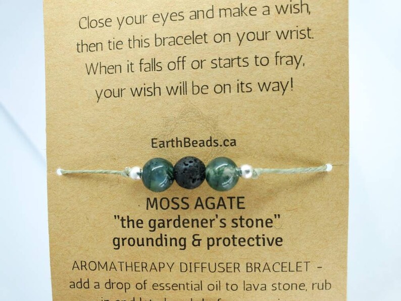 May include: A green moss agate stone bracelet with a black lava stone bead and silver spacer beads. The bracelet is tied with a light green string. The card reads "Close your eyes and make a wish, then tie this bracelet on your wrist. When it falls off or starts to fray, your wish will be on its way!"  "EarthBeads.ca"  "MOSS AGATE"  "the gardener's stone"  "grounding & protective"  "AROMATHERAPY DIFFUSER BRACELET - add a drop of essential oil to lava stone, rub"