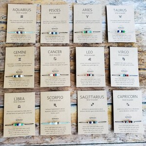 May include: Twelve cards with a brown background and white text. Each card features a zodiac sign symbol, name, and description. The cards are arranged in four rows of three. Each card has a wish bracelet with colourful beads and a string.